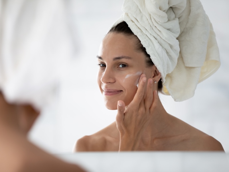 Happy cosmetic product consumer satisfied with moisturizing antiage cream. Young woman spreading nourishing lotion on facial skin after shower, smiling at reflection in mirror. Beauty care concept